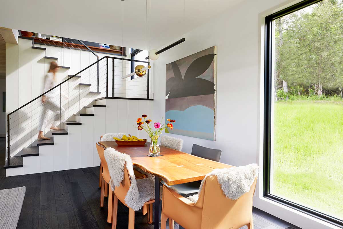 A person walking up a staircase from a minimalist, Scandinavian dining area next to a large Marvin window looking out to a lush lawn.