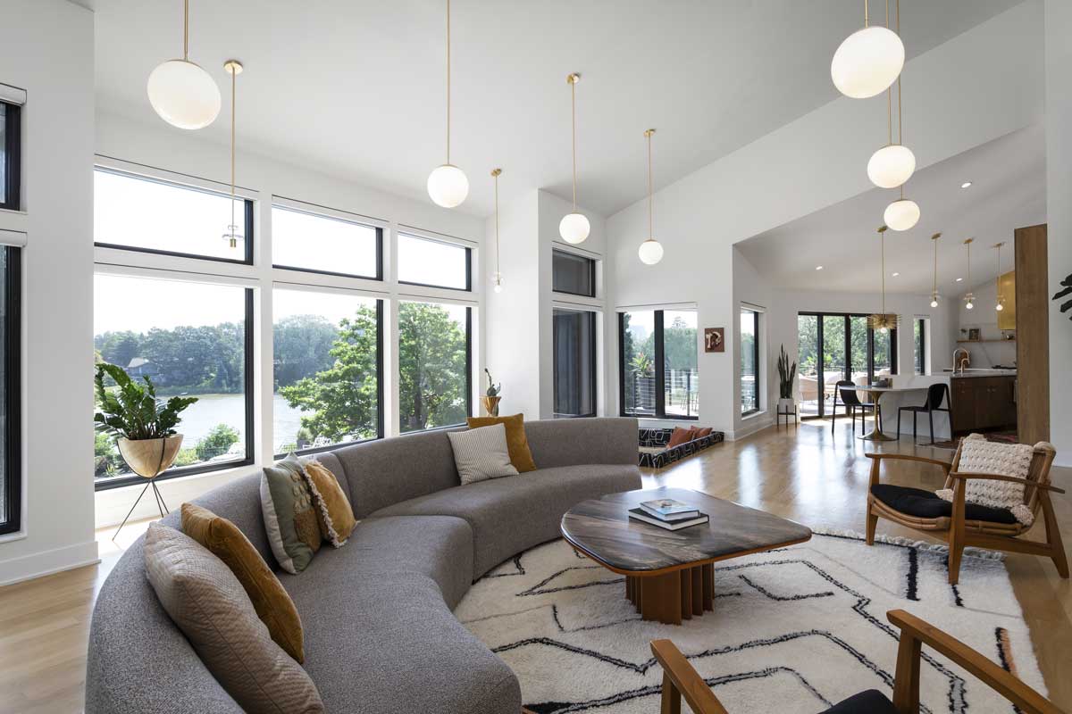 A modern living room featuring a curved gray couch with gold and white globe lights above and a view of a lake through Marvin Essential windows.