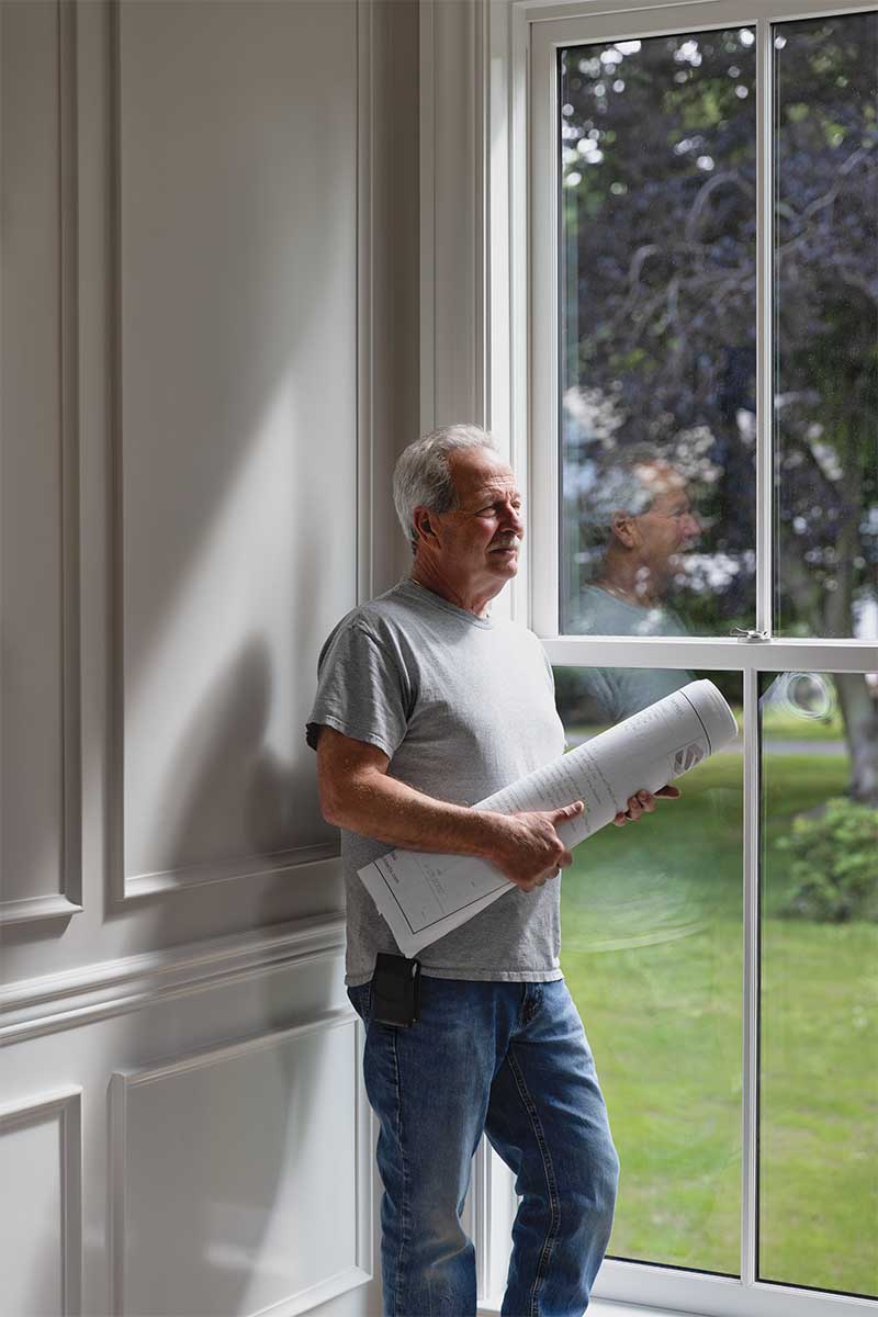 A portrait of builder Paul Jackson of Jackson's General Carpentry holding blueprints of Hadley House, next to a Marvin window inside the home.