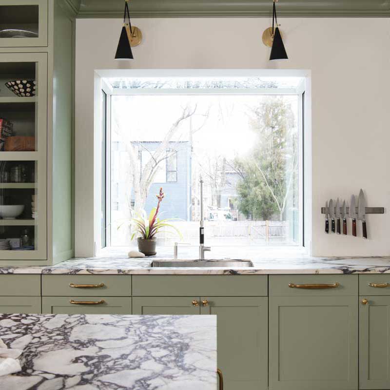 A contemporary kitchen with black and white marble countertops, sage green cabinets and a Marvin Skycove above the sink.