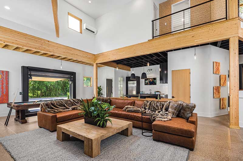 Hydraulic commercial garage door with cedar opens in a modern industrial-style home to reveal a recreation center with Marvin Ultimate Picture windows.