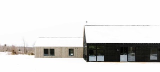 An image of a winter’s day outside of Svart Hus, a passive home in Door County, Wisconsin. Imagine two box-like buildings that are separate but connected. Roofs covered with snow. 