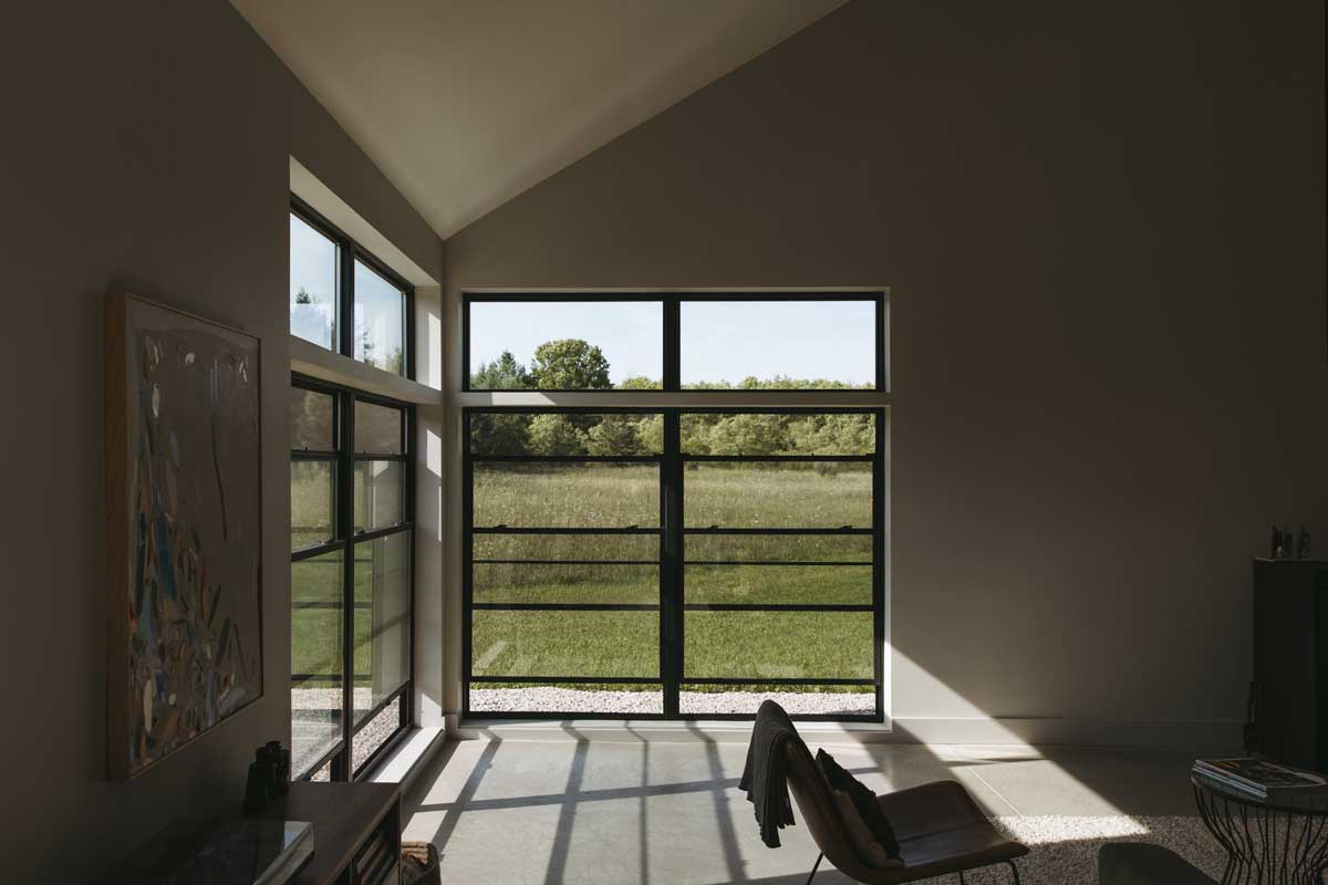 A modern, miniimalist living room with a wood-stove fireplace and a clean white walled interior, looking outward through black framed Marvin Essential windows to the snow covered forest beyond.